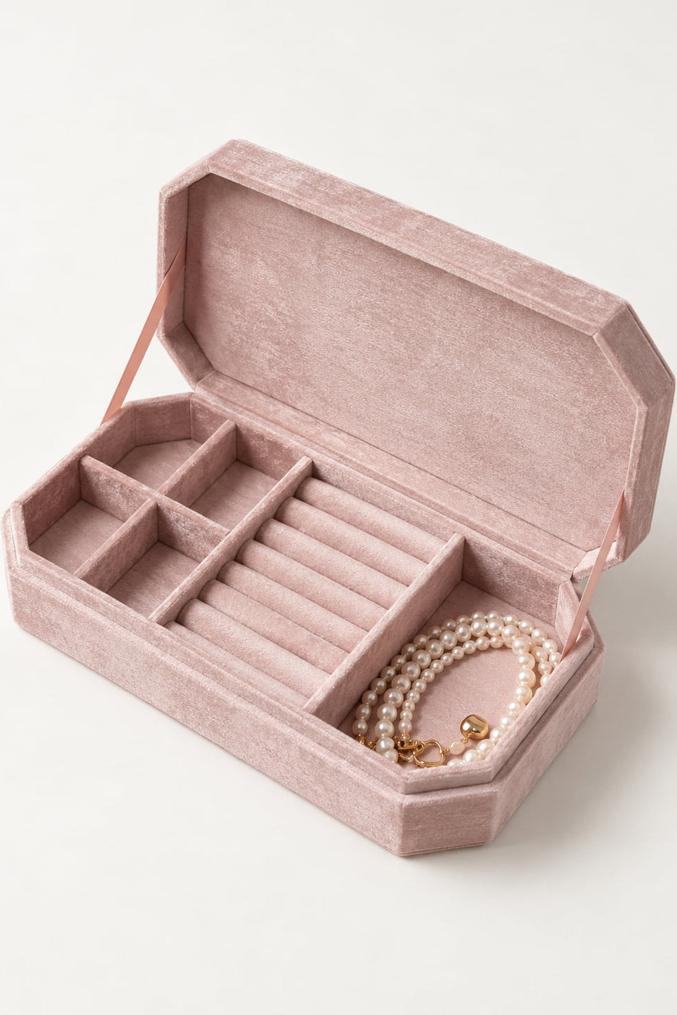 An open dusty pink velvet jewelry box with multiple compartments and a pearl necklace inside, photographed on a clean off-white background.