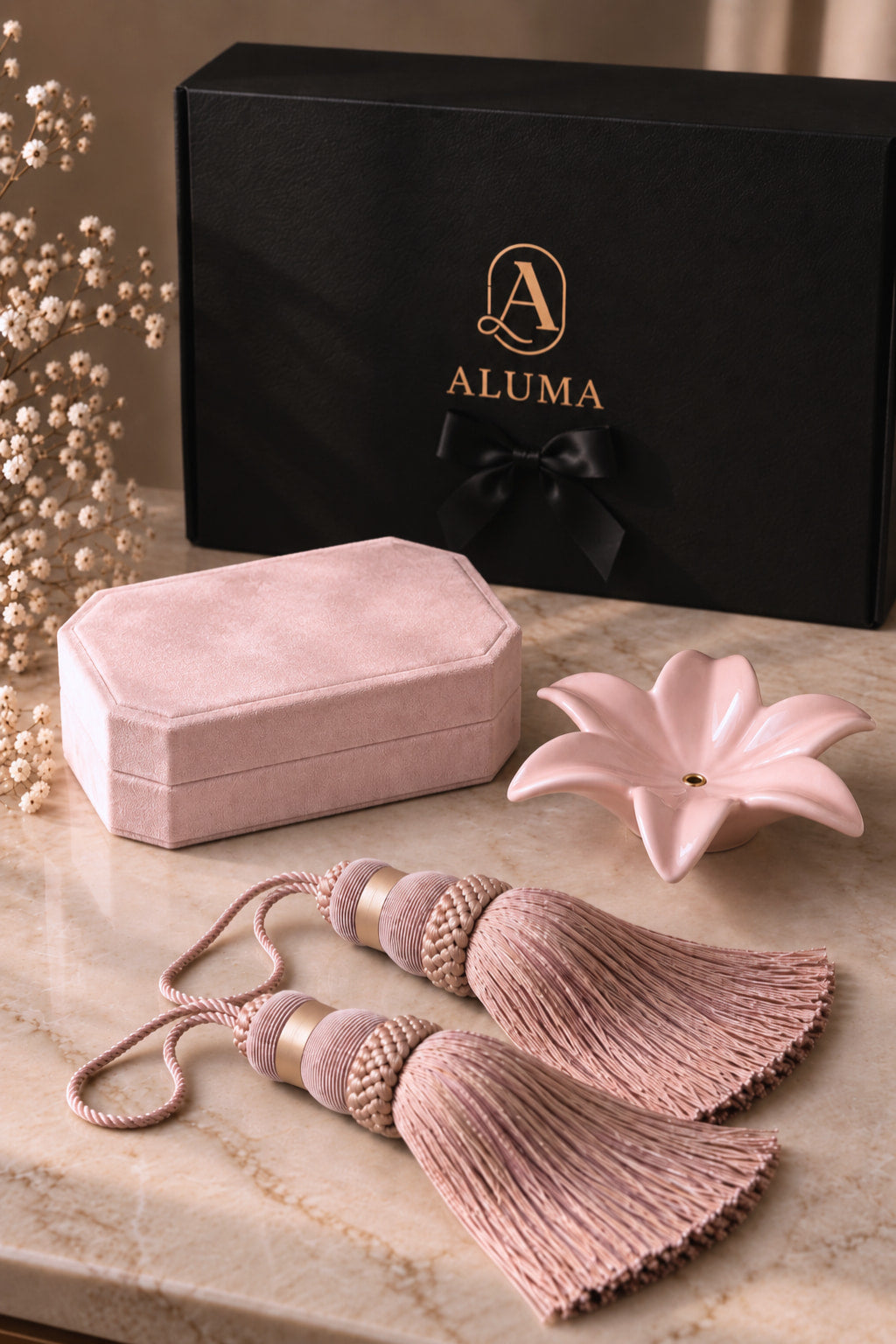 A luxury gift set featuring a dusty pink velvet jewelry box, matching tassel ties, a pink ceramic flower-shaped dish, and a black ALUMA branded gift box on a marble surface.