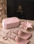 A luxury gift set featuring a dusty pink velvet jewelry box, matching tassel ties, a pink ceramic flower-shaped dish, and a black ALUMA branded gift box on a marble surface.