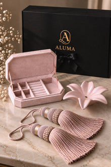 A pink velvet jewelry box shown open with its lid behind it, alongside a black ALUMA branded box, a pink flower-shaped dish, and two matching pink tassels on a marble surface.