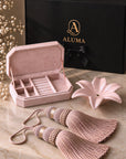 A pink velvet jewelry box shown open with its lid behind it, alongside a black ALUMA branded box, a pink flower-shaped dish, and two matching pink tassels on a marble surface.