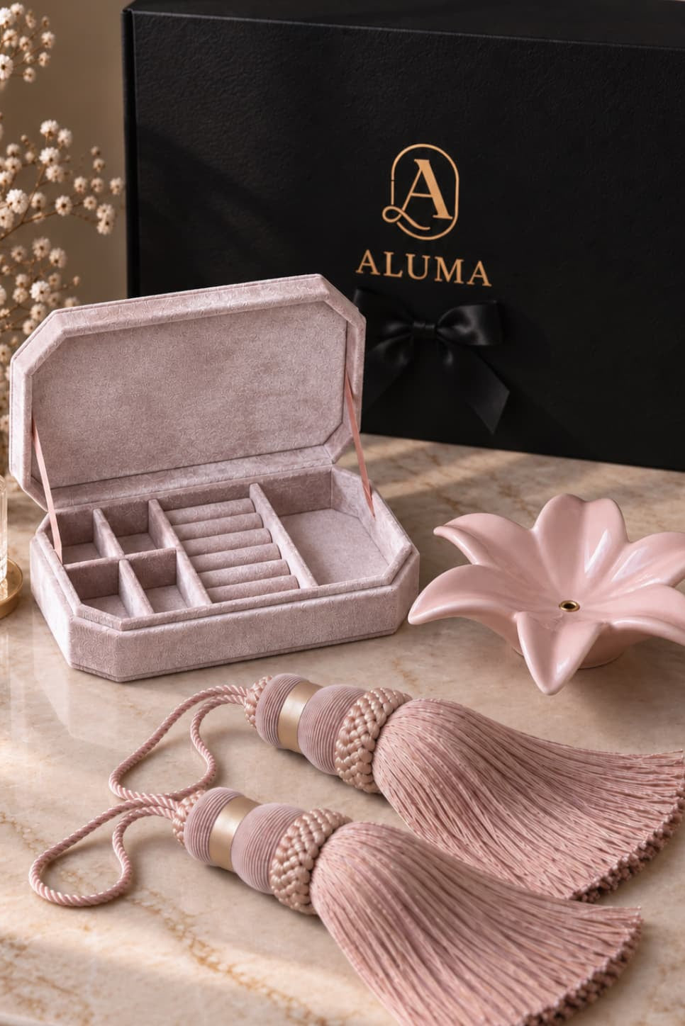A luxury jewelry organizer set by ALUMA, featuring a dusty pink velvet jewelry box with compartments, matching tassel ties, a pink lotus-shaped dish, and a black gift box with gold lettering, arranged on a beige marble surface.