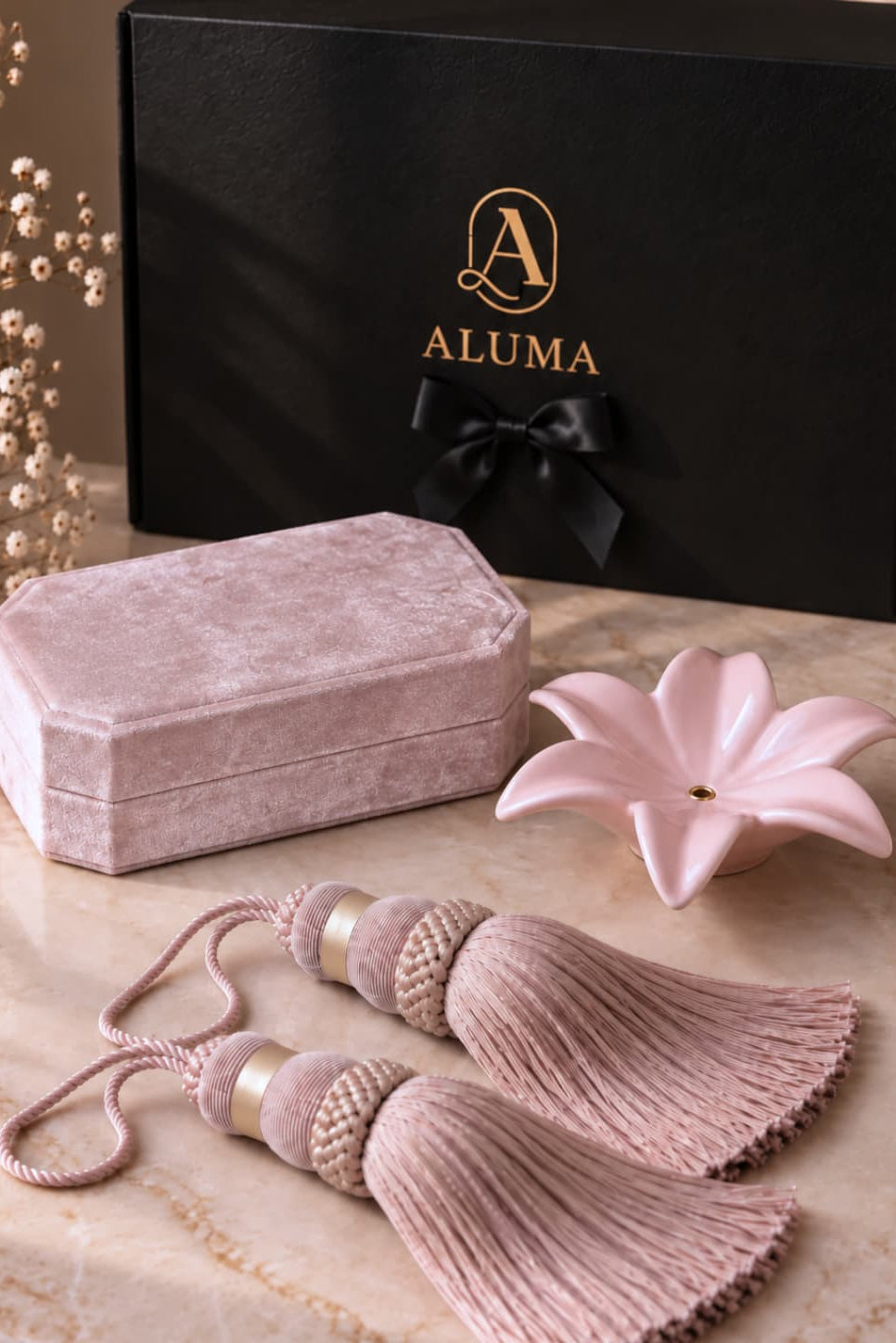 A luxury gift set featuring a dusty pink velvet box, two matching tassel ornaments with gold-toned metallic accents, a light pink ceramic flower-shaped dish, and a black gift box with gold 'ALUMA' branding.