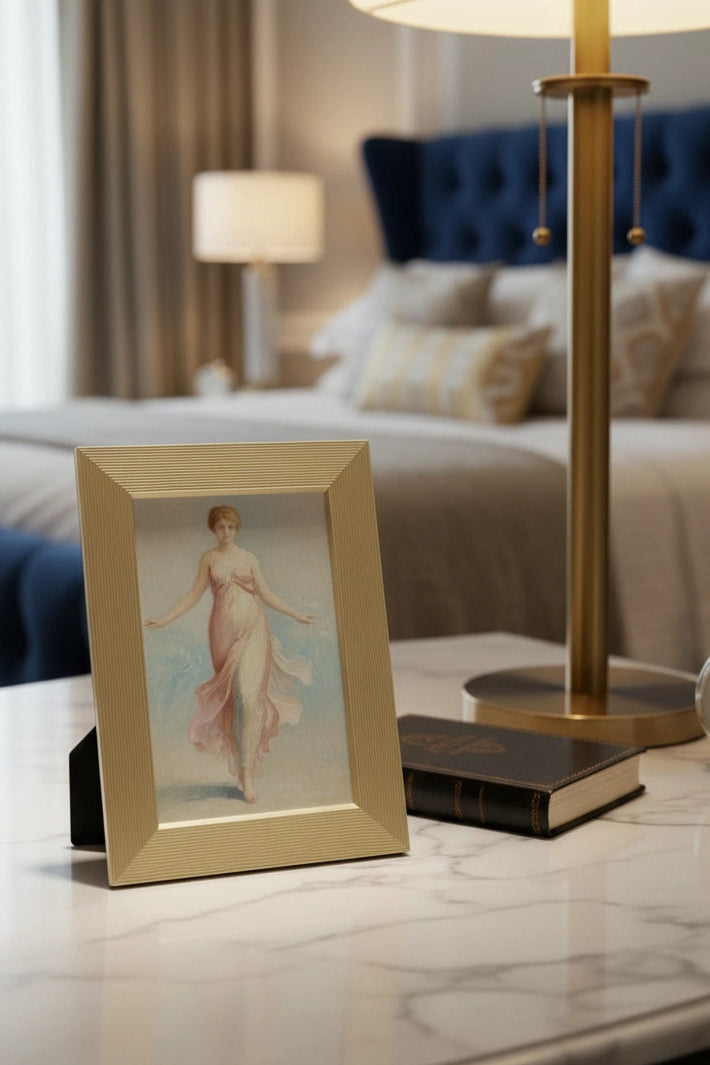 A luxury metal framed vintage-style illustration of a woman in a flowing dress sits on a marble bedside table in a bedroom, alongside a gold table lamp, a small book, and a white clock. The background shows a bed with a tufted blue headboard and soft neutral curtains.