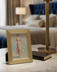 A luxury metal framed vintage-style illustration of a woman in a flowing dress sits on a marble bedside table in a bedroom, alongside a gold table lamp, a small book, and a white clock. The background shows a bed with a tufted blue headboard and soft neutral curtains.