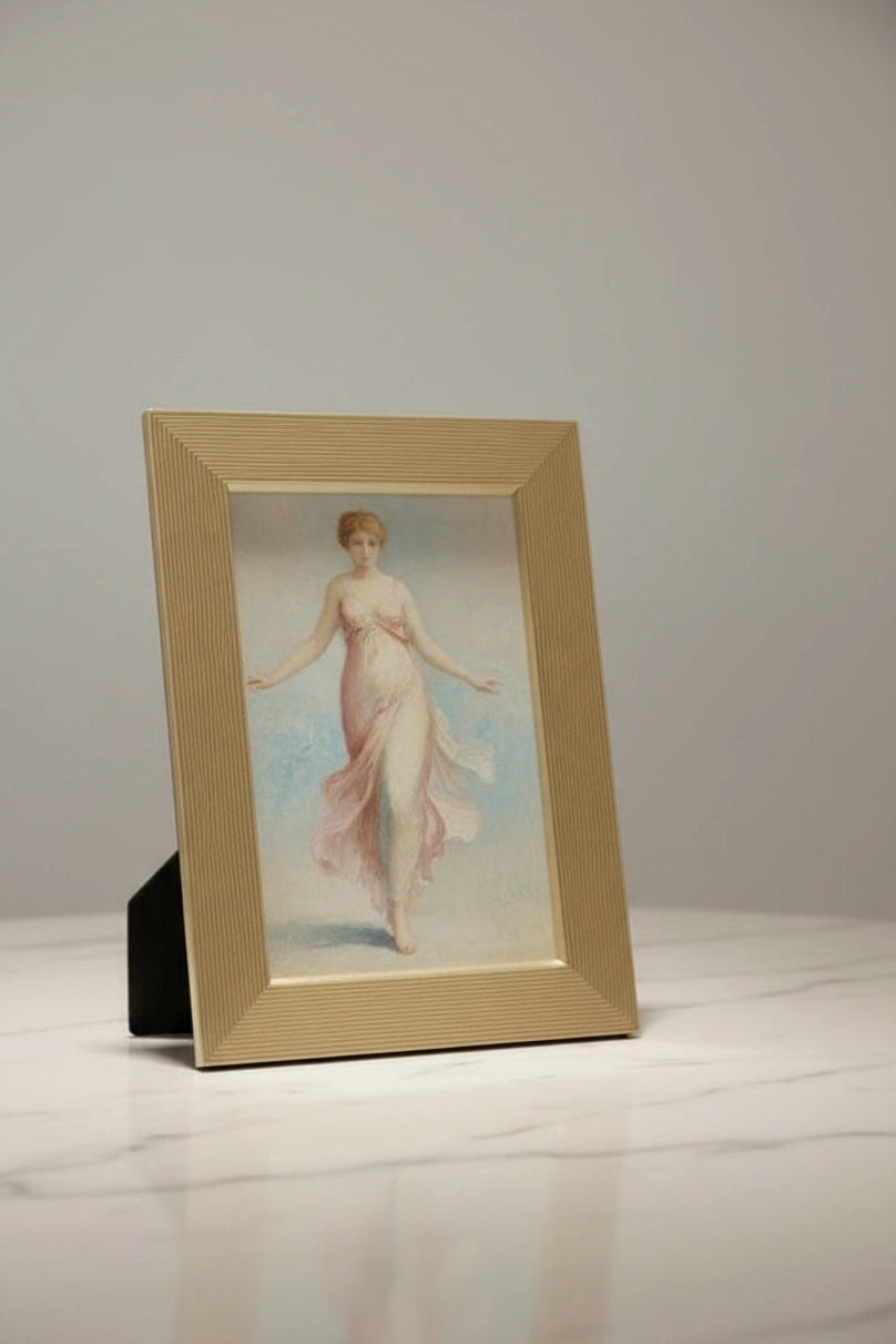 A luxury gold  framed vintage-style painting of a woman in a flowing pale pink dress, displayed on a white marble surface against a light gray wall.