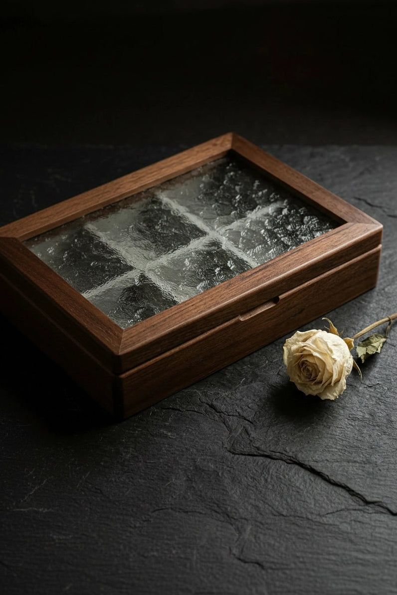 A rectangular wooden keepsake box with a textured glass window, shown on a dark slate surface next to a dried cream-colored rose.