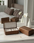 A wooden jewelry box set displayed on a white marble surface in a softly lit bedroom setting, with one open box showing compartments and a necklace inside, plus matching closed boxes and a mirror reflecting the room.