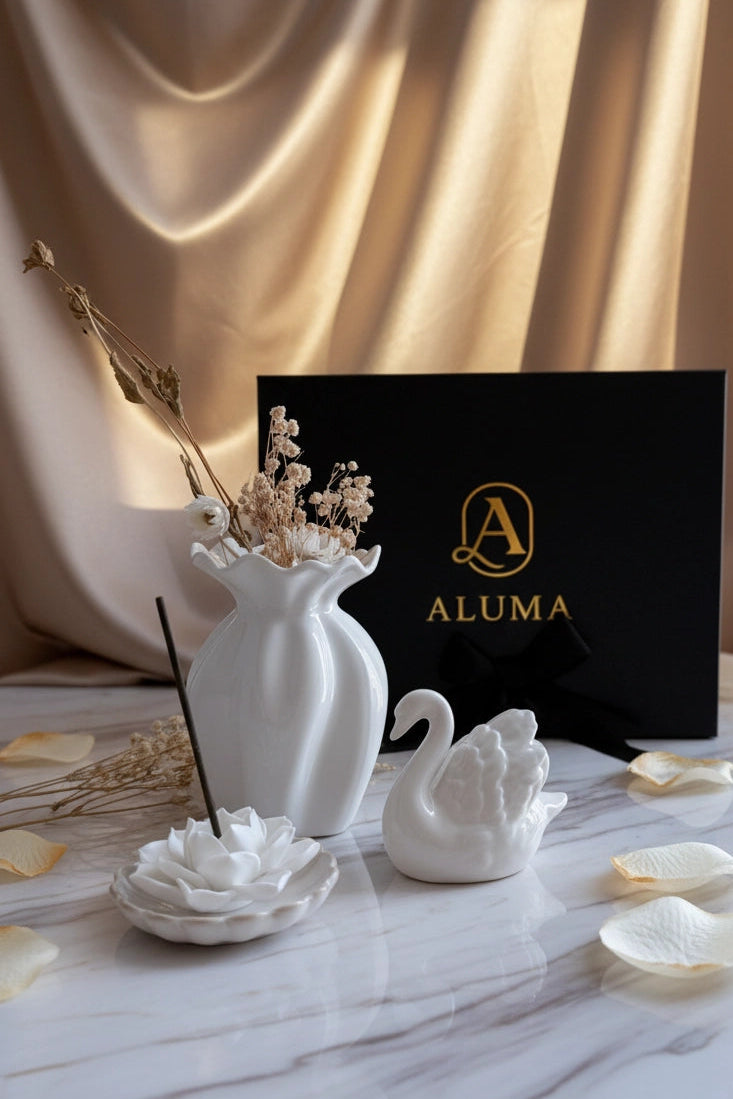 A styled product display featuring a white ceramic diffuser vase with dried botanicals, a white swan figurine, a white flower-shaped incense holder with a black incense stick, and a black ALUMA branded box, arranged on a marble surface with gold satin drapery in the background.