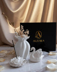A styled product display featuring a white ceramic diffuser vase with dried botanicals, a white swan figurine, a white flower-shaped incense holder with a black incense stick, and a black ALUMA branded box, arranged on a marble surface with gold satin drapery in the background.