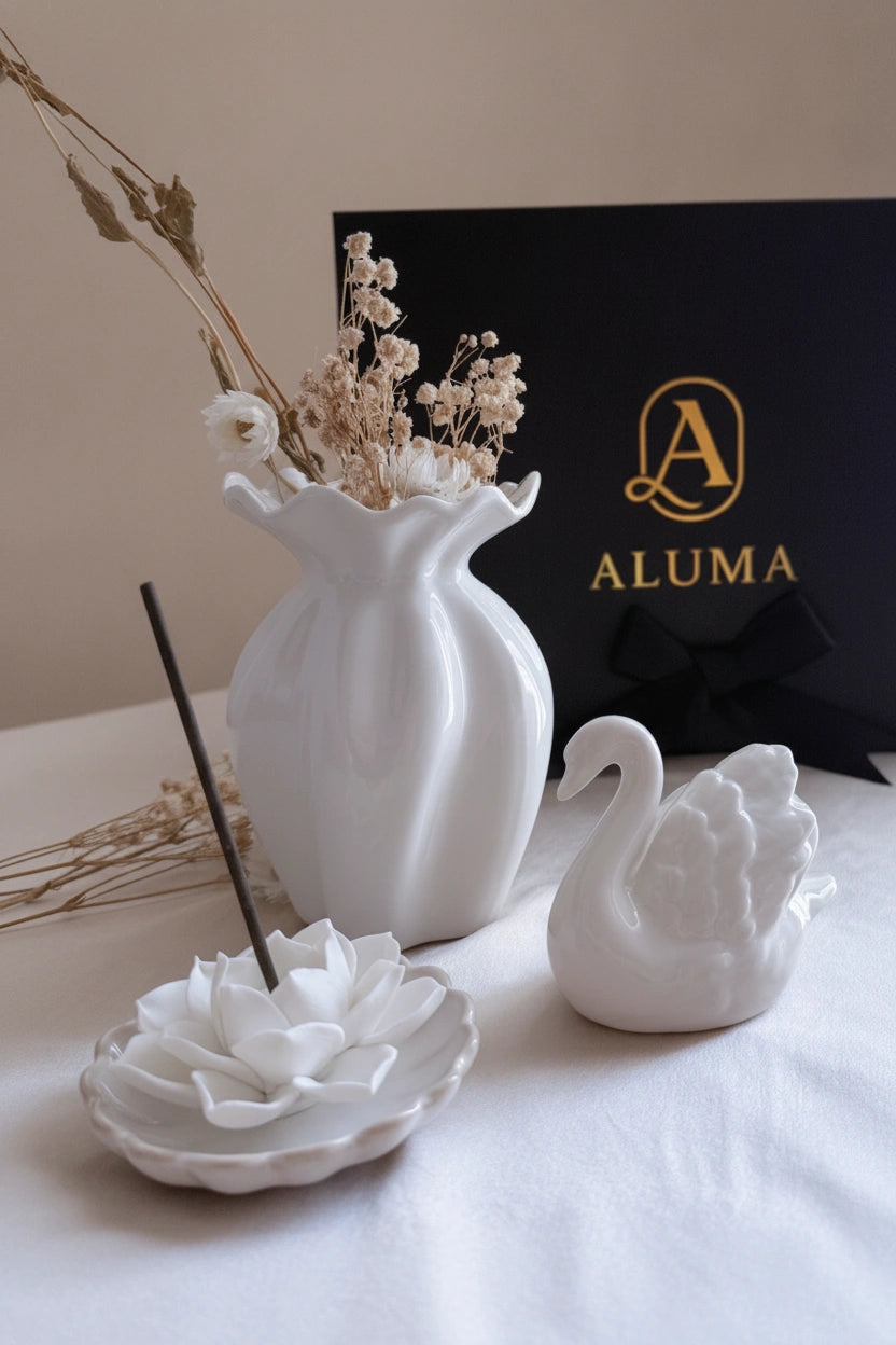 White ceramic home decor set including a vase with dried flowers, a swan figurine, a lotus-shaped incense holder with a black incense stick, and a black ALUMA gift box in the background.