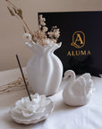 White ceramic home decor set including a vase with dried flowers, a swan figurine, a lotus-shaped incense holder with a black incense stick, and a black ALUMA gift box in the background.