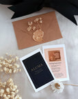 A styled flat-lay of wedding stationery on a light fabric background, featuring a kraft paper envelope with a gold wax seal, a black invitation card with the name ALUMA, a pink invitation card with wedding details, and dried white flowers.