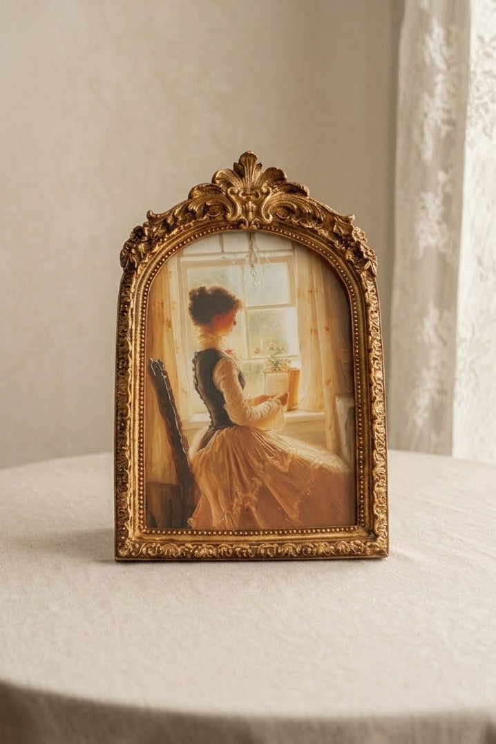 An ornate gold-framed vintage-style picture frame displaying a painting of a woman in a yellow dress by a window, set on a round table against a neutral cream wall and lace curtain background.