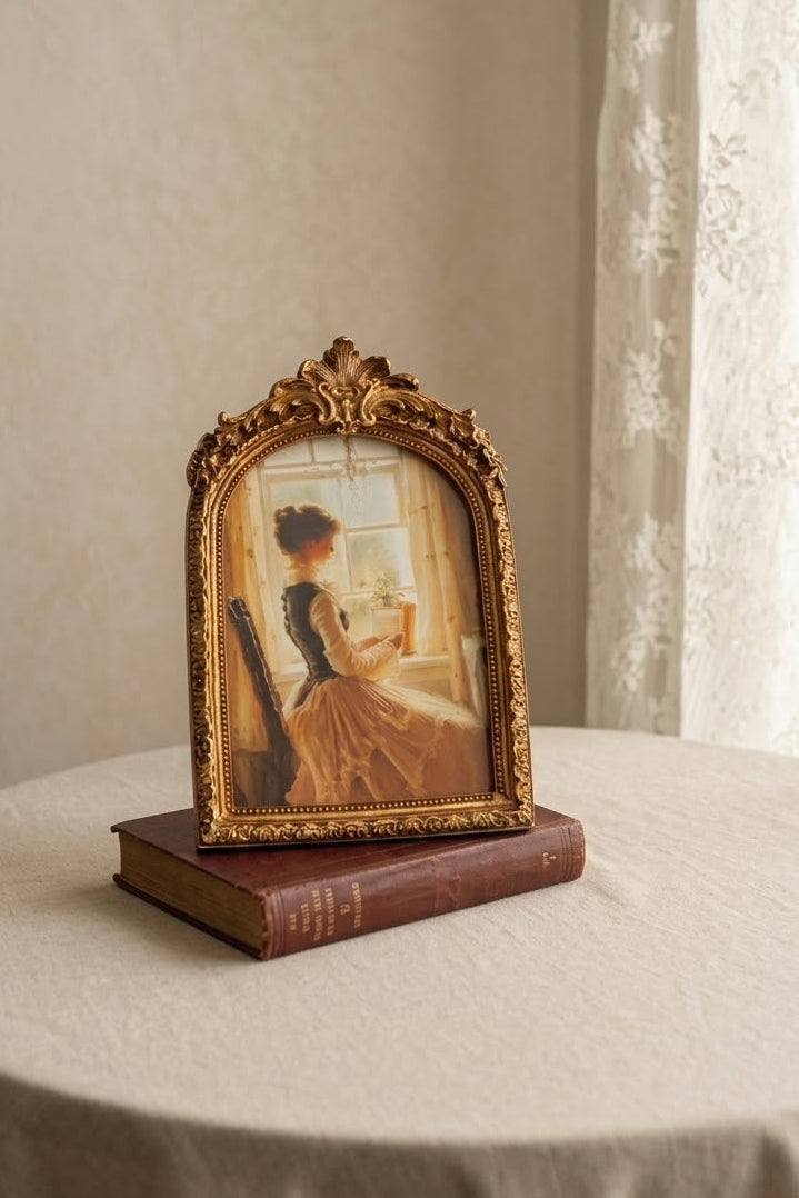 An ornate gold-framed vintage art print depicting a woman in a dress by a window, displayed on top of a brown book on a beige tablecloth next to sheer curtains.