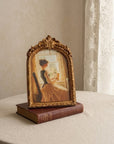 An ornate gold-framed vintage art print depicting a woman in a dress by a window, displayed on top of a brown book on a beige tablecloth next to sheer curtains.