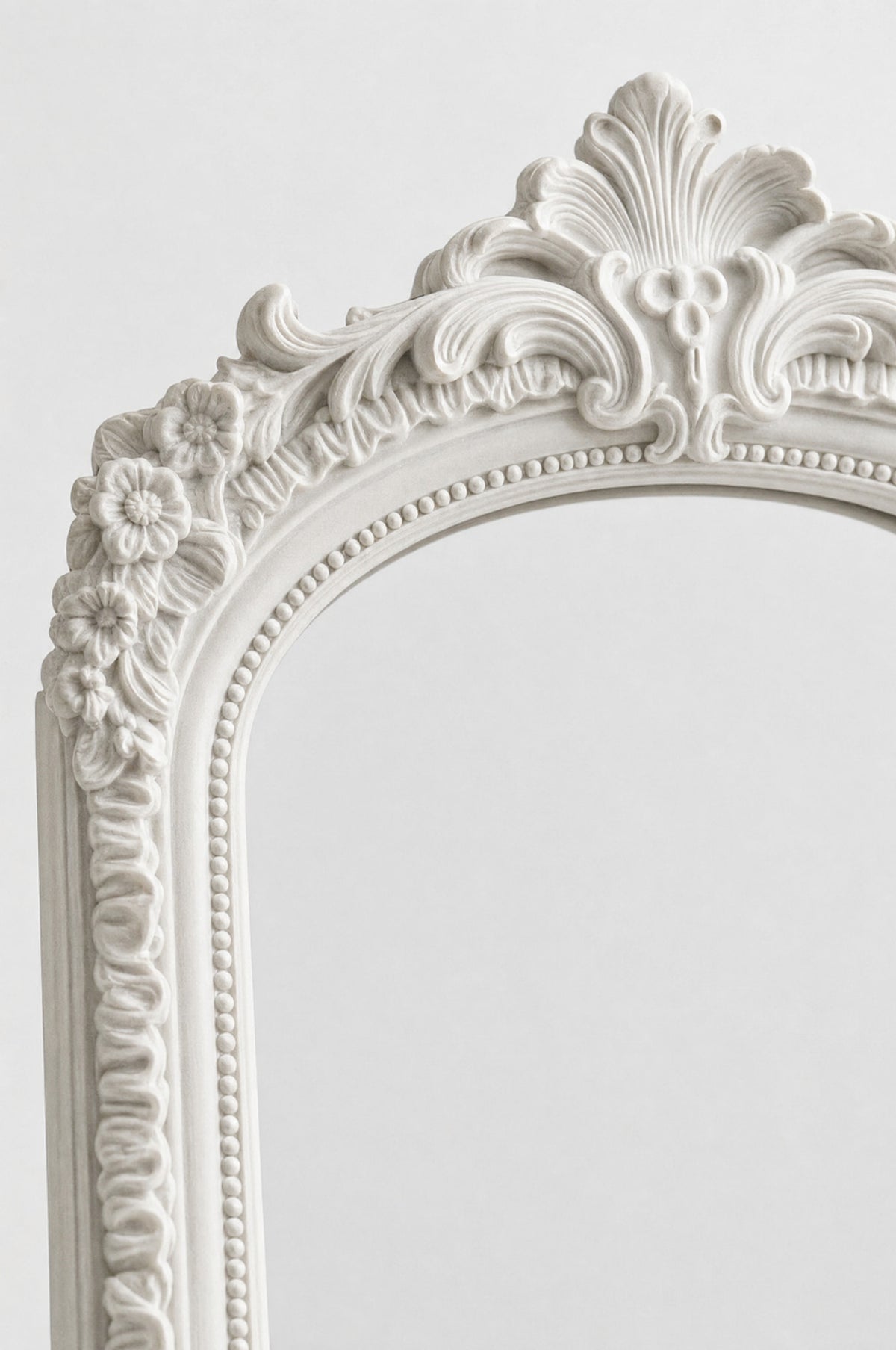 Close-up of an ornate white decorative mirror frame with intricate floral and scrollwork details against a light gray background.