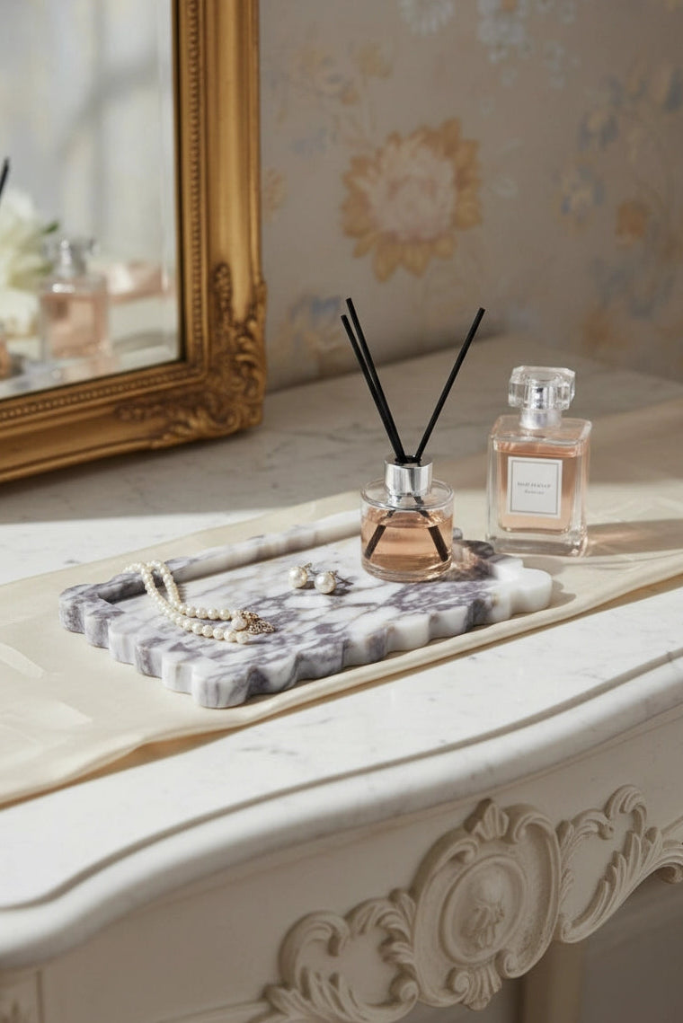A styled interior scene showing a marble tray holding a reed diffuser and a small perfume bottle on an ornate cream console table, with a gold-framed mirror and floral wallpaper in the background.