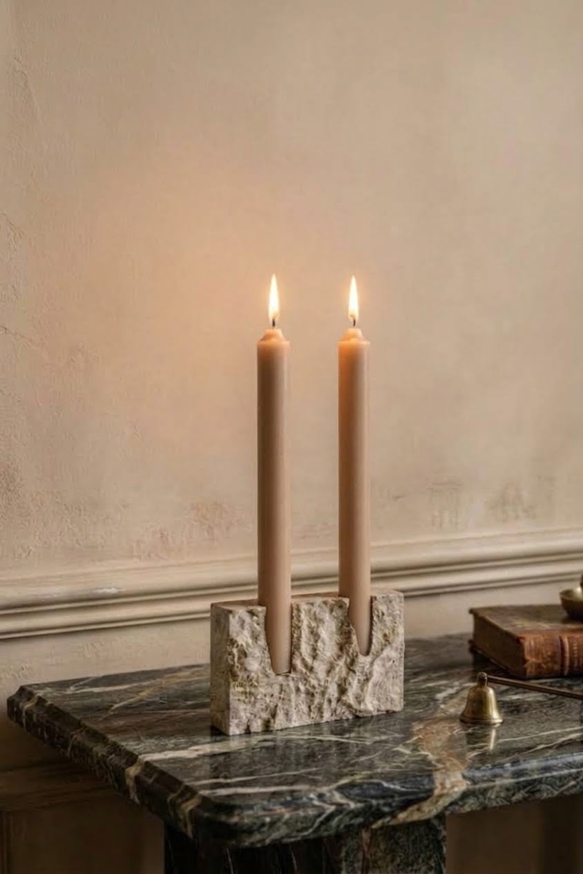 A two-tiered candle holder made of textured beige stone, holding two lit beige taper candles, set on a dark marble table against a textured beige wall.
