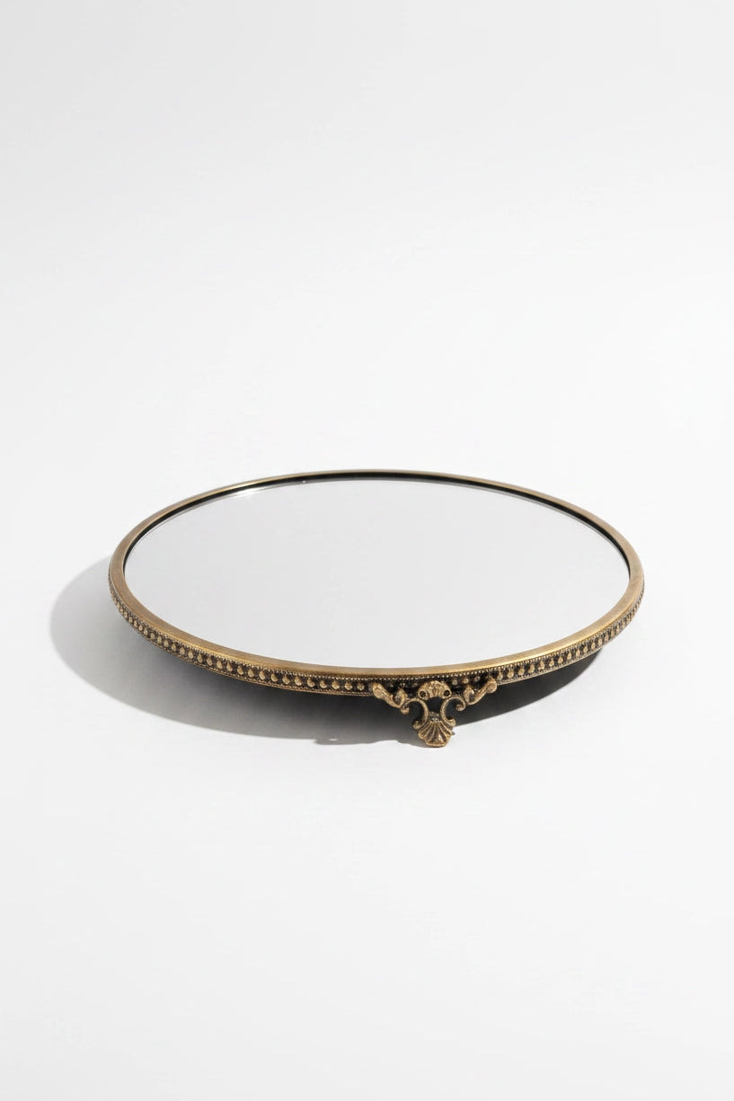 A round decorative mirror with an ornate antique gold brass tray , featuring a beaded border and a small central scroll detail, set against a plain white background.