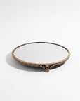 A round decorative mirror with an ornate antique gold brass tray , featuring a beaded border and a small central scroll detail, set against a plain white background.