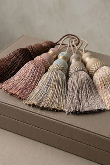 Five decorative tassels in earthy neutral tones including brown, dusty rose, beige, and cream, arranged on top of a textured taupe leather box.