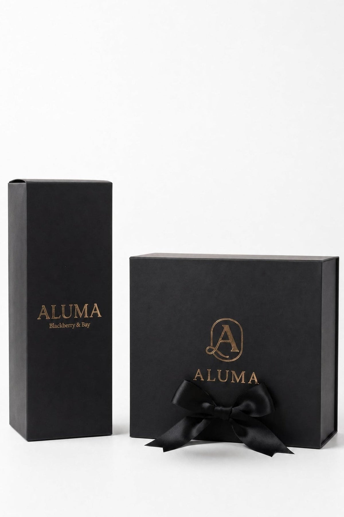 Two black ALUMA fragrance boxes on a white background, one tall rectangular box and one square gift box with a black ribbon bow, both featuring gold lettering.