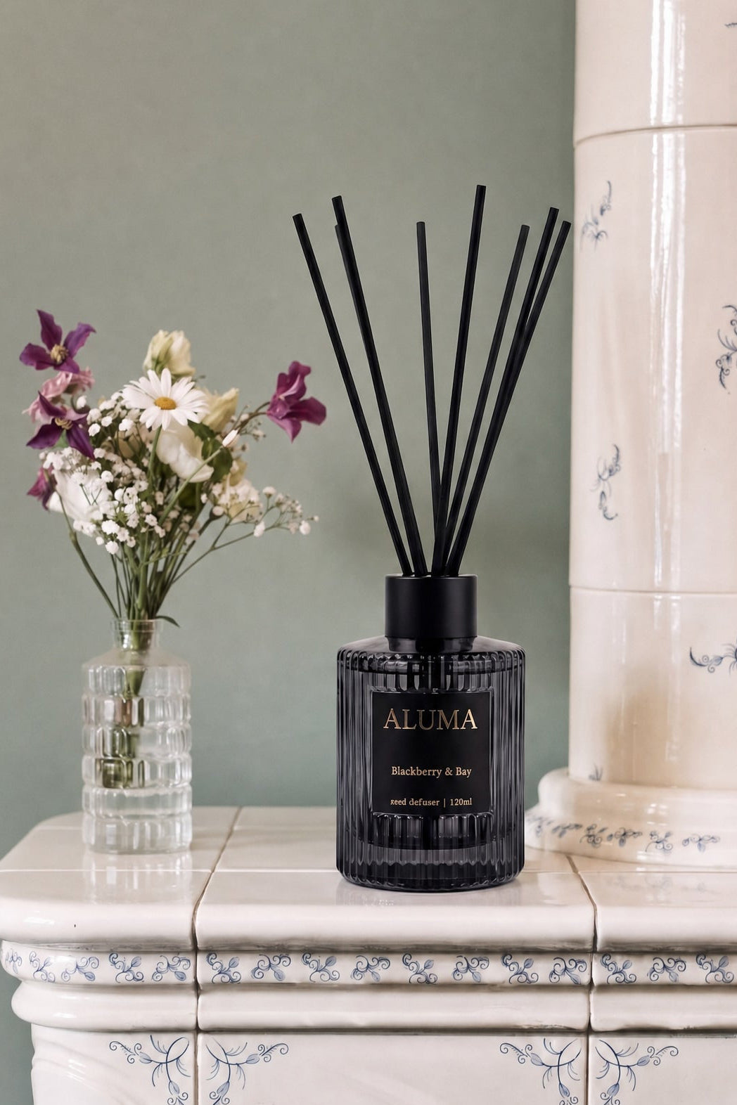 A reed diffuser bottle with black reeds sits on a white tiled console table next to a small vase of flowers, with a tall cream-colored ceramic column in the background.