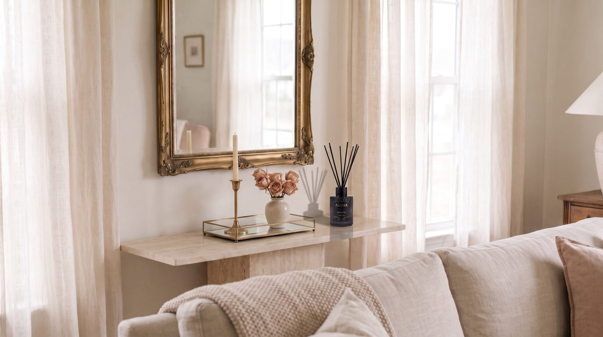 A styled interior scene with a beige sofa in the foreground, a console table beneath a large ornate gold mirror, sheer cream curtains, and a reed diffuser on the marble table.