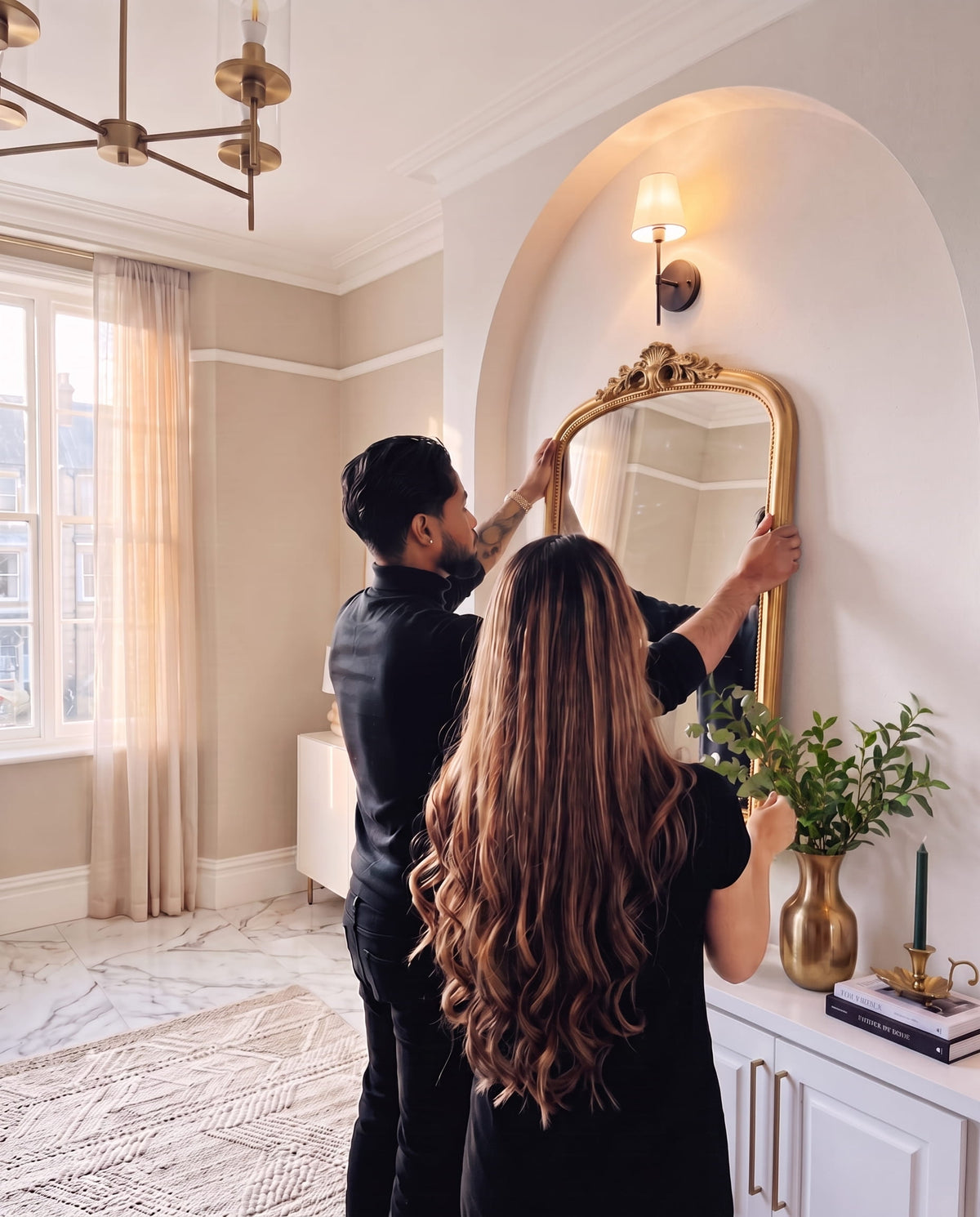 A man and a woman founders of Aluma Home in black outfits are shown from behind as they adjust a large, ornate gold-framed mirror on a white arched wall in a beautifully decorated room with beige walls, a marble floor, and a white console table.