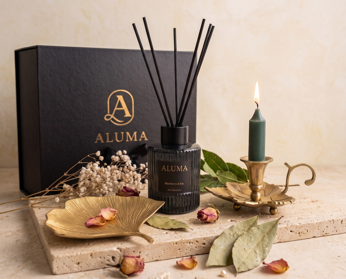 ALUMA reed diffuser bottle with black reeds, a black gift box with gold logo, a teal candle in a brass leaf holder, dried flowers, leaves, and rose petals on a stone slab against a warm beige background.