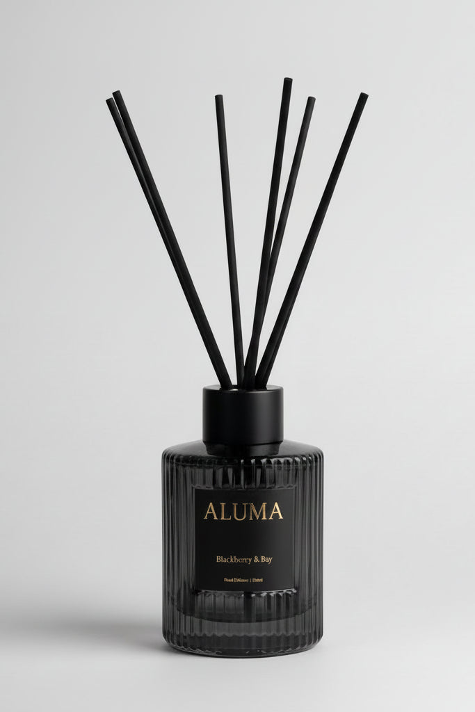 A reed diffuser bottle with black reeds, a ribbed black glass body, and a black label with gold lettering reading 'ALUMA' and 'Blackberry & Bay' on a white background.