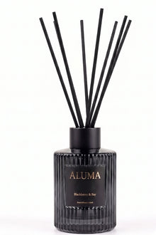 A reed diffuser bottle with black reeds, a ribbed black glass body, and a black label with gold lettering reading 'ALUMA' and 'Blackberry & Bay' on a white background.
