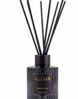 A reed diffuser bottle with black reeds, a ribbed black glass body, and a black label with gold lettering reading 'ALUMA' and 'Blackberry & Bay' on a white background.