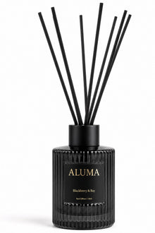 A reed diffuser bottle in black ribbed glass with black reeds, featuring a black label with gold lettering that reads 'ALUMA' and 'Blackberry & Bay'.