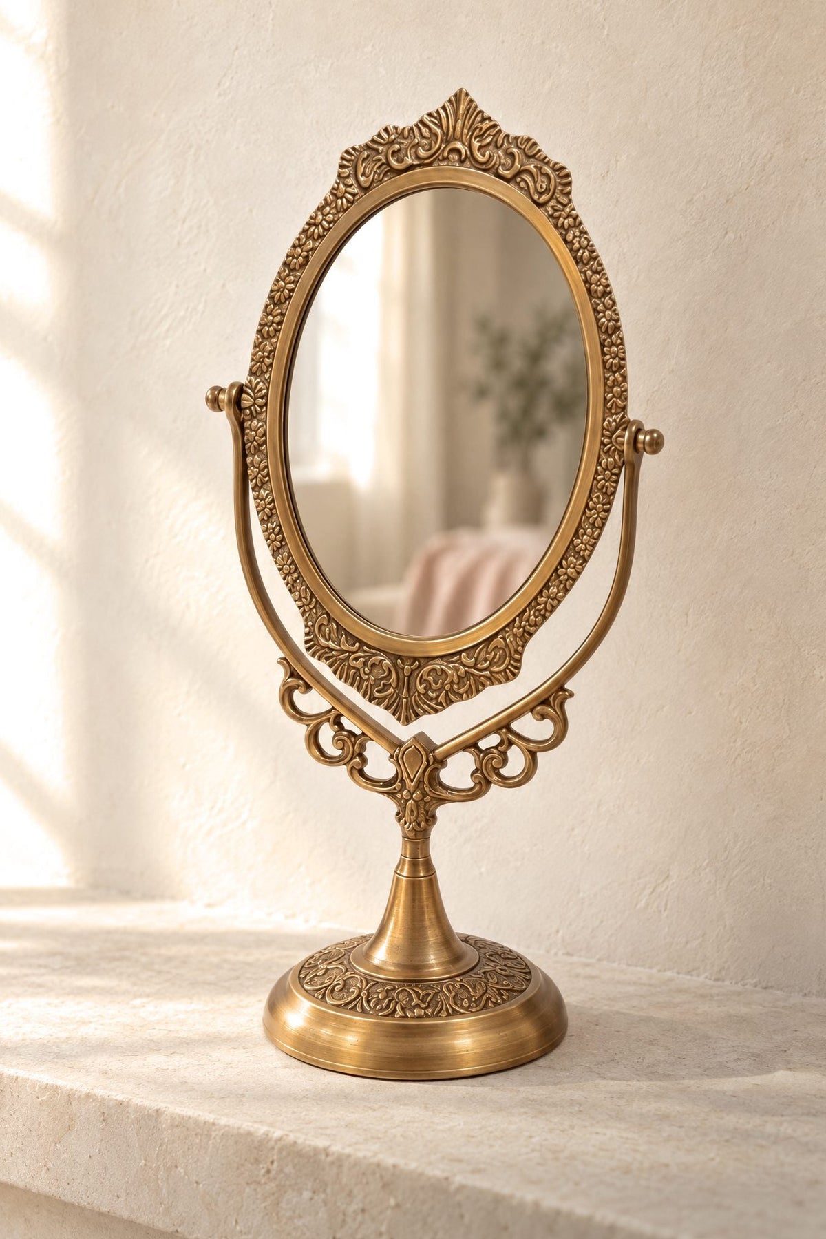 An ornate brass gold vanity mirror with a decorative scrollwork frame and pedestal base, set on a light stone surface against a textured cream wall.