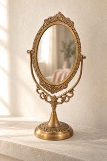 An ornate brass gold vanity mirror with a decorative scrollwork frame and pedestal base, set on a light stone surface against a textured cream wall.