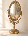 An ornate brass gold vanity mirror with a decorative scrollwork frame and pedestal base, set on a light stone surface against a textured cream wall.