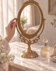 An ornate brass gold-toned oval vanity mirror with a decorative frame and stand, displayed on a marble-top dresser in a softly lit bedroom setting. and hand touching the mirror 