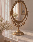 An ornate  brass gold vanity mirror with a decorative oval frame and matching pedestal base, styled on a marble surface in a softly lit room.