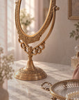 An ornate brass gold-toned freestanding oval mirror with intricate scrollwork, displayed on a marble surface in a softly lit interior setting.