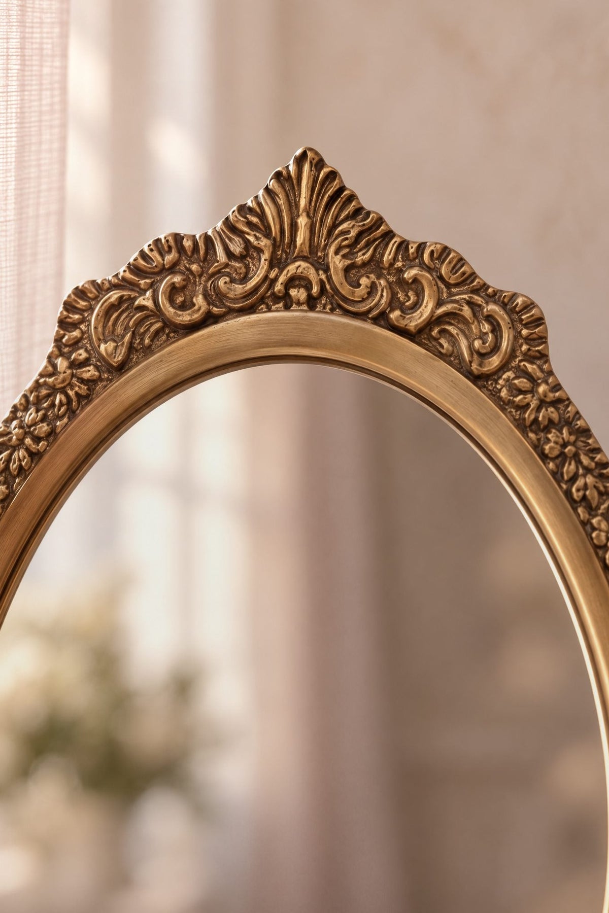Close-up of an ornate oval brass mirror with a carved gold frame, set against a soft beige curtain background.