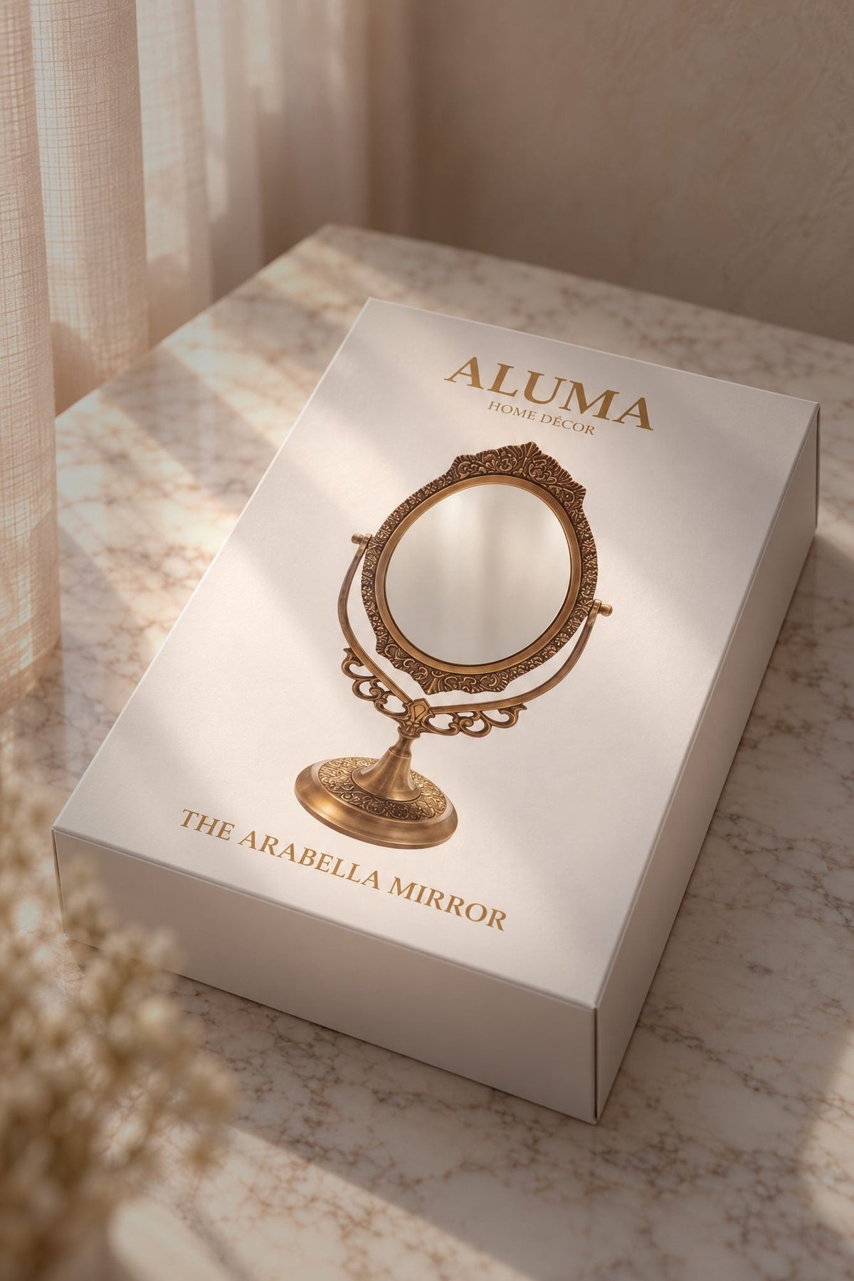 A white rectangular product box for ALUMA 'The Arabella Mirror' sits on a marble-like surface with warm sunlight and soft curtains in the background. The box features a gold illustration of an ornate vanity mirror.