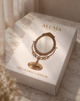 A white rectangular product box for ALUMA 'The Arabella Mirror' sits on a marble-like surface with warm sunlight and soft curtains in the background. The box features a gold illustration of an ornate vanity mirror.