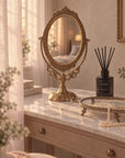 An ornate brass gold-toned oval vanity mirror with a decorative frame and stand, displayed on a marble-top dresser in a softly lit bedroom setting.