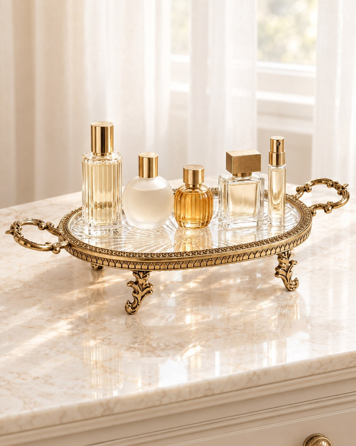An ornate gold metal tray with a clear glass top sits on a cream marble dresser in front of a softly lit window with white curtains. Several perfume and cosmetic bottles are arranged on the tray.