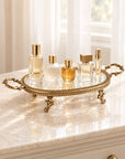 An ornate gold metal tray with a clear glass top sits on a cream marble dresser in front of a softly lit window with white curtains. Several perfume and cosmetic bottles are arranged on the tray.
