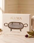 A cream product box labeled 'ALUMA HOME DECOR ATHENA TRAY' stands on a light marble surface in a softly lit interior. The front shows a cutout window revealing a silver tray inside, with decorative pearls, greenery, and berries placed around it.