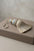 A decorative tassel with a cream fringe, a braided beige-and-white center, a light blue cylindrical section, and a small gold accent, resting on a taupe leather box against a neutral beige background.