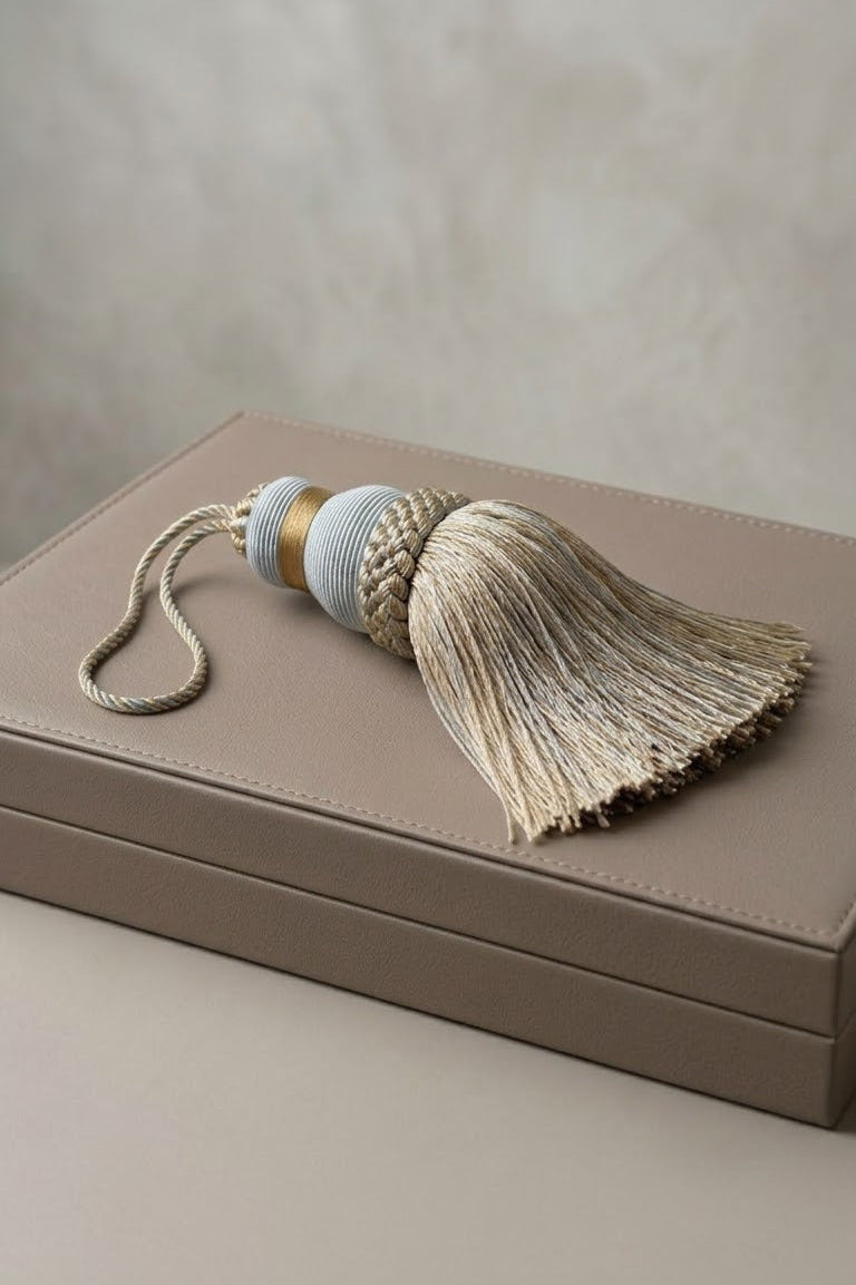A decorative tassel with a cream fringe, a braided beige-and-white center, a light blue cylindrical section, and a small gold accent, resting on a taupe leather box against a neutral beige background.